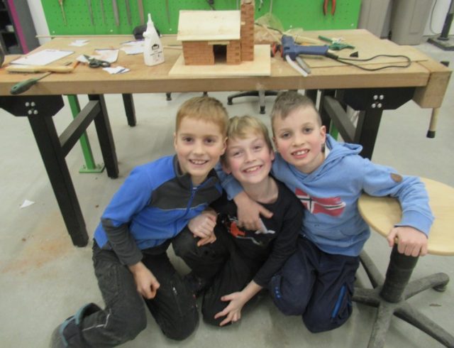 Drei Kinder sitzen vor einer Werkbank. Auf der Werkbank steht ein selbstgebautes Miniaturhaus mit Kamin aus Holz und Ziegeln.