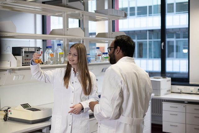 zwei junge Erwachsene in Laborkitteln betrachten eine orangene Flüssigkeit in einem Kolben