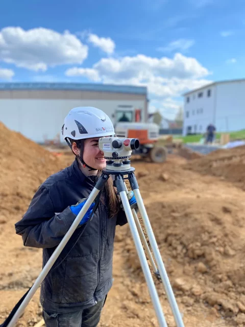 junge Frau mit Helm auf einer Baustelle schaut durch Vermessungsgerät