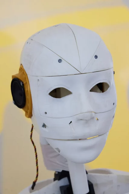 Weiße Roboterkopfnachbildung mit gelben Ohren vor gelbem Hintergrund.