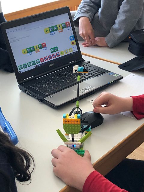 ein Roboter aus Bausteinen neben einem Laptop mit der zugehörigen Programmierungssoftware in einfacher Darstellung