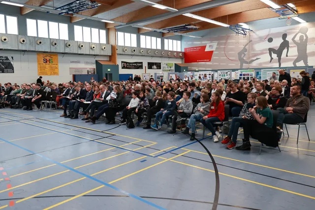 viele Menschen sitzen in Sporthalle auf Stühlen