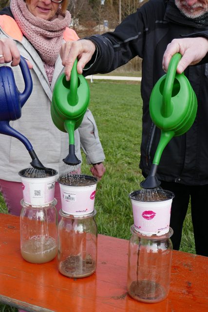 drei ältere Menschen gießen einen kleinen Blumentopf, der Erde enthält und auf einem Einmachglas steht, in dem sich Wasser sammelt
