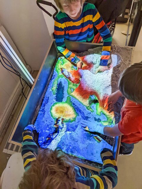 drei Kinder modellieren eine Landschaft mit Hilfe von Sand und einer projezierten Höhenkarte