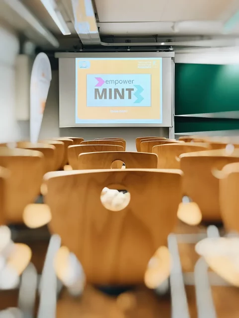 leere Holzstühle vor Leinwand mit Logo empower MINT