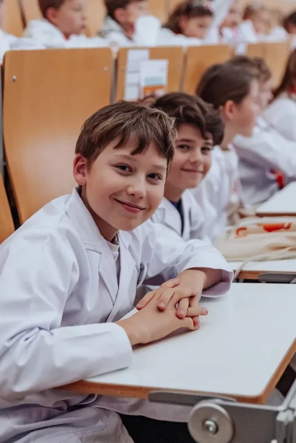 Kinder in Laborkitteln sitzen lächelnd in einem Hörsaal