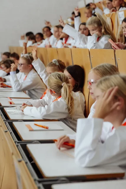 Kinder in Laborkitteln sitzen in Hörsaal und melden sich