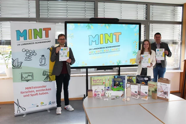 Drei Personen halten MINT-Adventskalender und stehen vor einem Bildschirm.