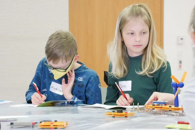 zwei Kinder arbeiten in einem Heft, vor ihnen steht ein kleines Modellwindrad