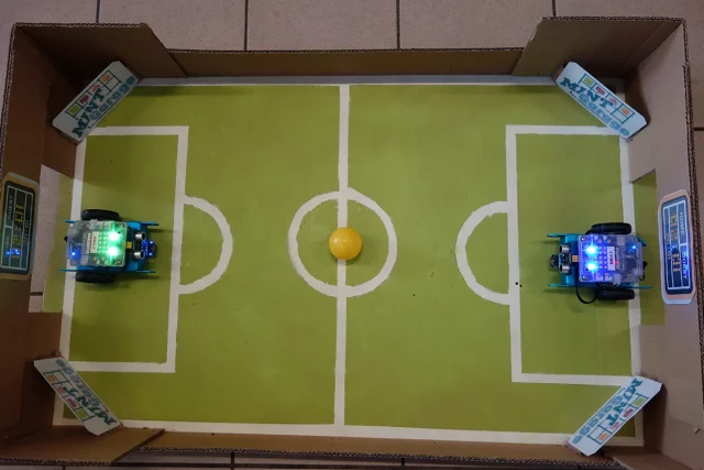 kleines Fußballfeld aus Pappe, in den Toren stehen zwei Robotor mit Rädern, bereit loszufahren, im Mittelkreis liegt ein gelber Ball