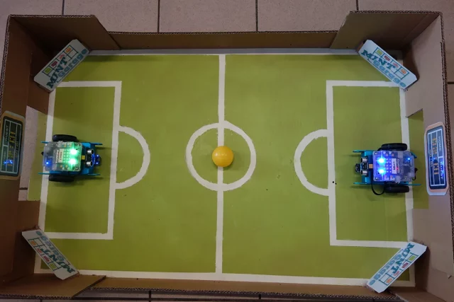 kleines Fußballfeld aus Pappe, in den Toren stehen. Zwei Robotor mit Rädern, bereit loszufahren, im Mittelkreis liegt ein gelber Ball