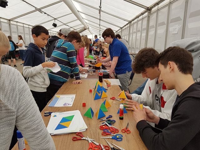 viele junge Menschen stehen an Tischen und basteln kleine pyramidenförmige Körper mithilfe von Vorlagen