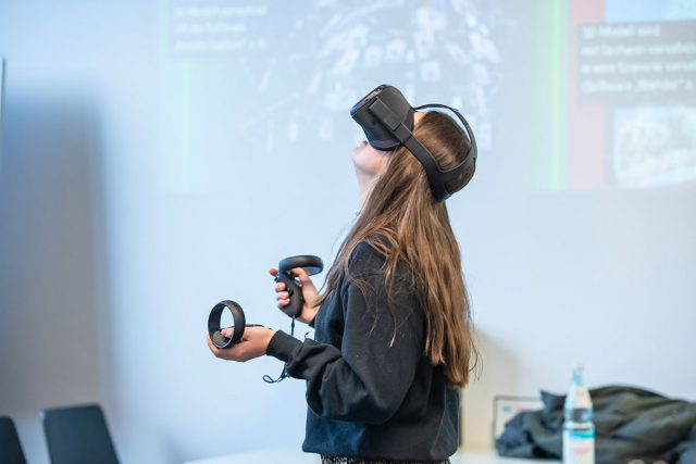 junge Frau mit VR-Brille auf dem Kopf und Controllern in der Hand, schaut nach oben