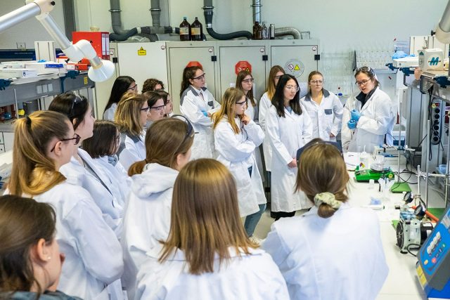 viele junge Frauen in Laborkitteln stehen in einem Halbkreis um einen Labortisch