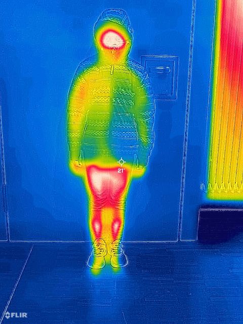 Aufnahme einer Wärmebildkamera, eine Person mit Winterjacke ist zu sehen, das meiste ist gelb und blau, Beine und Gesicht sind rot und weiß