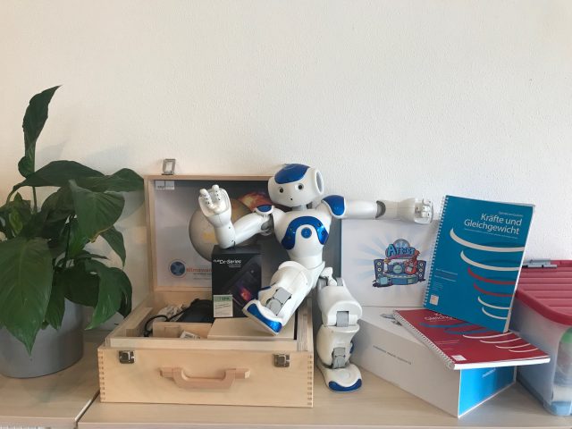 kleine Roboter in menschenähnlicher Form sitzt auf Holzkiste, neben ihm stehen physikalische Hefte