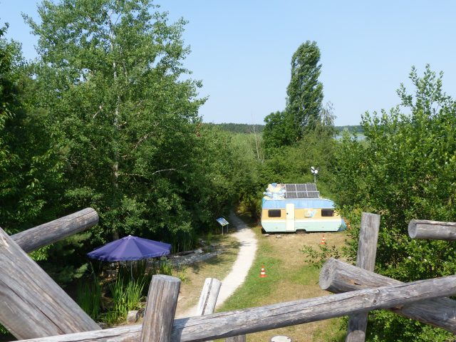 Bunter Camper mit Solarpanels neben einem Sonnenschirm in einer grünerLandschaft.