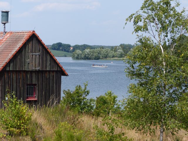 Haus am Seeufer mit vorbeifahrendem Boot und Bäumen.
