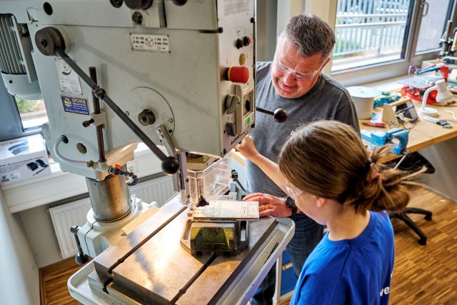 Person zeigt Kind den Umgang mit einer Metallbohrmaschine.