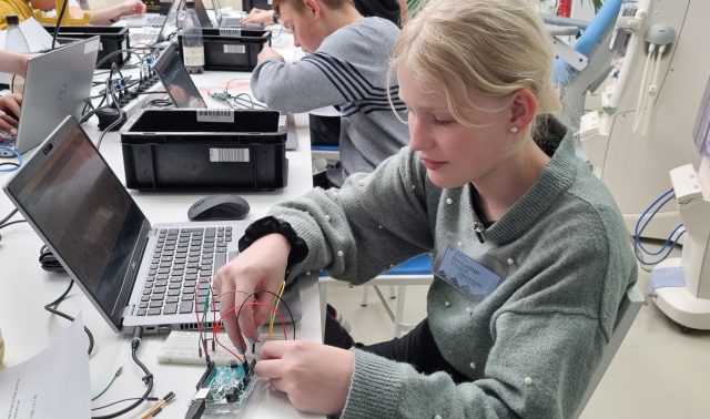 Schülerin arbeitet an technischer Schaltung mit Laptop im Hintergrund.