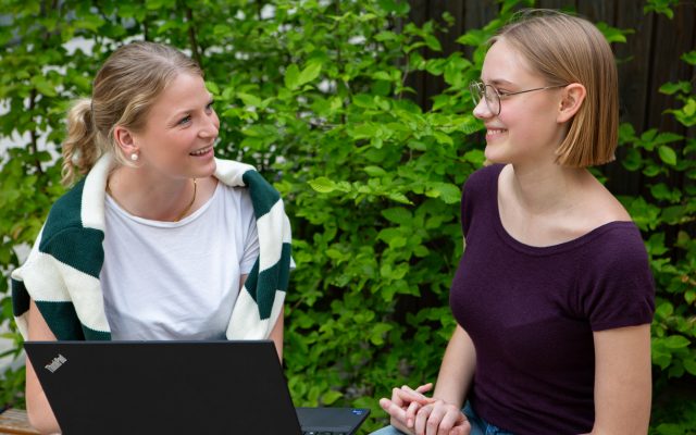 Zwei Frauen im Gespräch im Freien neben einem Laptop.