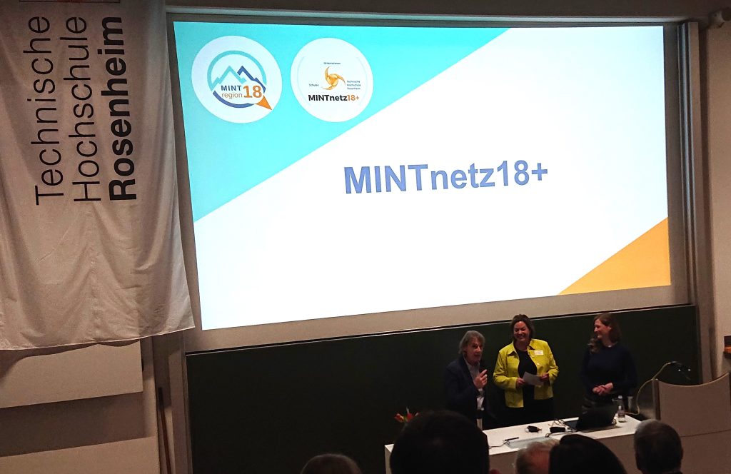 In einem Hörsaal stehen drei Frauen vor einer riesigen Leinwand. Auf der Leinwand steht MINTnetz18+. Oben links in der Ecke sind die Logos der MINTregion18 und des Netzwerkes MINTregion18+