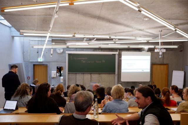 Ein Hörsaal mit Betonwänden und moderner Beleuchtung ist mit zahlreichen Personen besetzt, die an einem Netzwerktreffen teilnehmen. Die Menschen sitzen auf Holzbänken und unterhalten sich, während vorne an der Tafel „1. Netzwerktreffen MINT-Cluster Region 10“ geschrieben steht. Daneben ist eine Leinwand mit einer Präsentation zu sehen, auf der ebenfalls „1. Networking Meeting“ steht. Im Vordergrund sind Teilnehmer im Gespräch, einige haben Wasserflaschen oder Laptops vor sich. Die Atmosphäre wirkt lebendig und interaktiv.