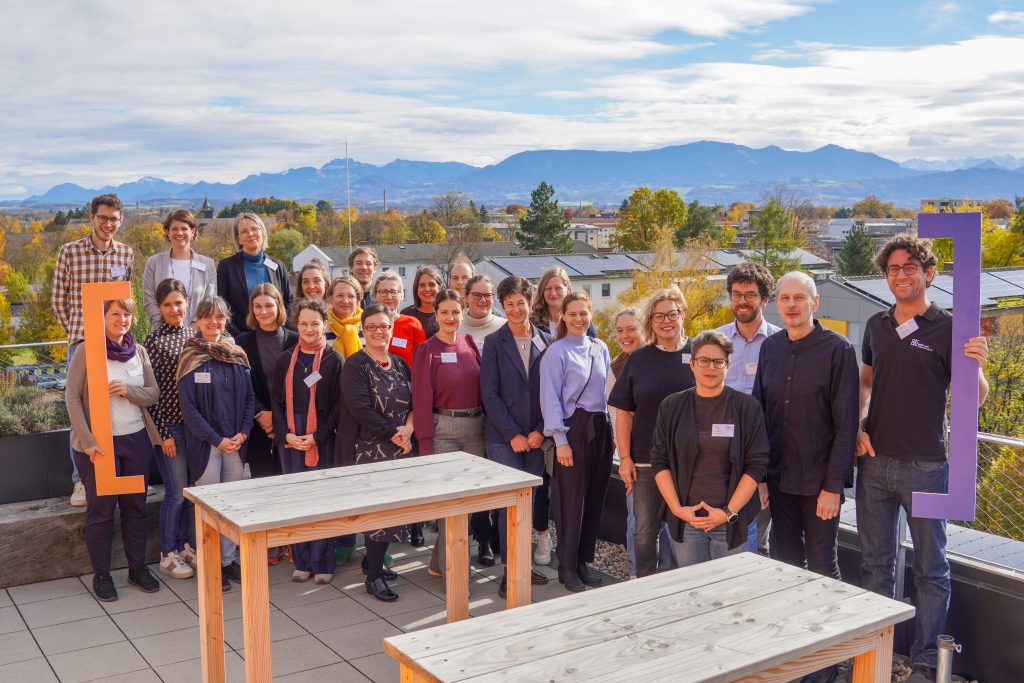 Teilnehmer und Teilnehmerinnen des Netzwerktreffens vom MINTraum bayern stehen für das Gruppenfoto auf der Dachterasse des Veranstaltungsgebäude. Im Hintergrund sind die Alpen zu erkennen.
