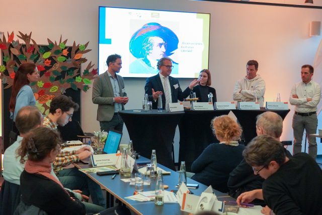 Teilnehmerinnen und Teilnehmer der Gesprächsrunde beim Netzwerktreffen stehen an drei Stehtischen. Im Hintergrund ist ein großer Bildschirm mit einem Bild von Goethe zu sehen.
