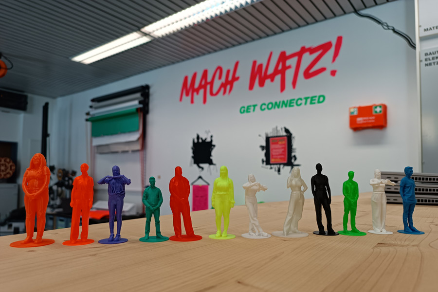 Personen aus dem 3D-Drucker in unterschiedlichen Farben stehen auf einem Holztisch. Im Hintergrund ist der Schriftzug Mach Watz! auf einer weißen Wald gemalt.