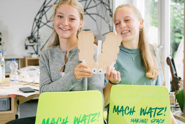 Zwei Mädchen sitzen auf Stühlen halten jeweils eine Holzarbeit hoch.